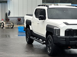 White 2025+ vehicle with Kia Tasman side steps, black steel side bars for strength and style, parked in a wet urban setting.